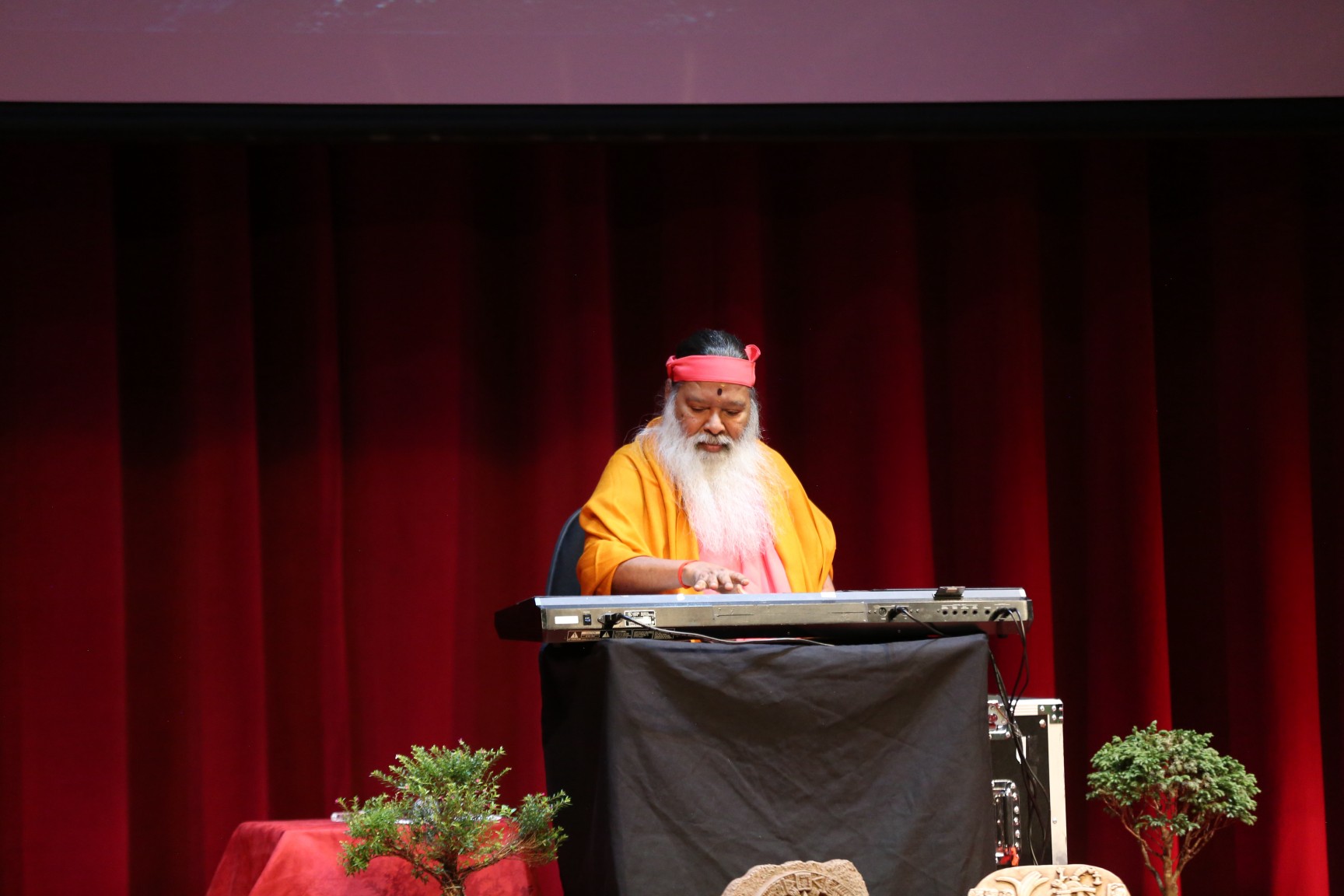 Sri Swamiji at keyboard — red curtain stage